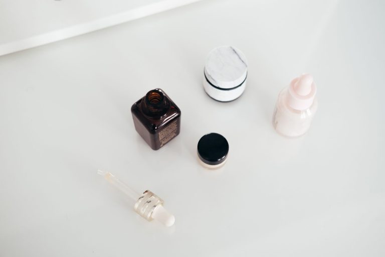 Otwarty flakon serum z pipetą obok słoiczków kremu na białym blacie